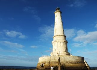 Le Phare de Corduan A white stone lighthouse nestled on a round three storey round base rises into a deep blue september sky.Tourists are mingling around the lighthouse and base which is accesible because of low tide.