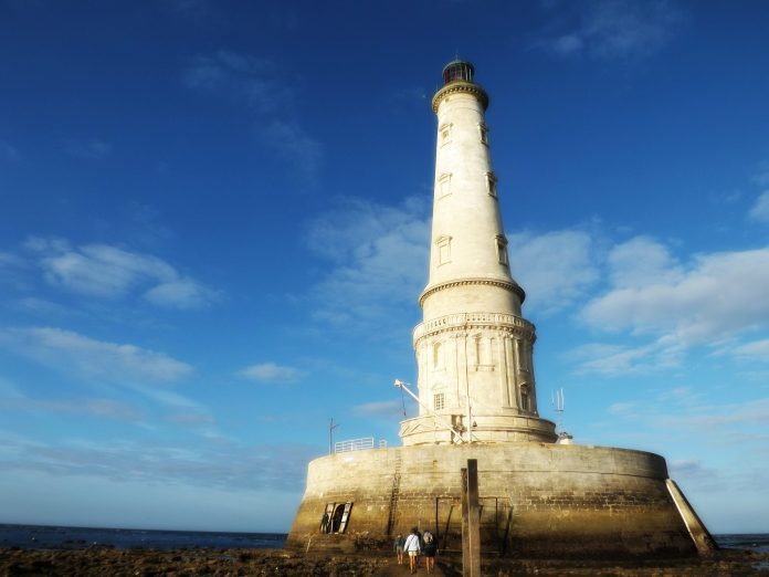 Un phare en pierre blanche niché sur une base ronde de trois étages s'élève dans un ciel bleu profond de septembre. Les touristes se mêlent autour du phare et de la base qui est accessible en raison de la marée basse.