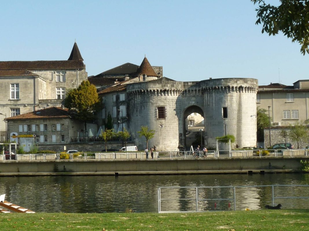 Une porte ronde en pierre avec deux tours cylindriques et une entrée voûtée se dresse au bord d'une rivière calme à Cognac ; des bâtiments historiques avec des toits en tuiles sont à l'arrière-plan, et quelques piétons marchent le long du chemin de la rivière sous un ciel dégagé.