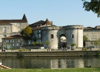Percer les secrets de la tapisserie culturelle du Cognac Une porte historique en pierre avec deux tours rondes et une entrée voûtée se dresse au bord du fleuve à Cognac, entourée de bâtiments anciens. Les gens se promènent à pied ou à vélo le long du chemin qui longe le fleuve, se faufilant dans la tapisserie culturelle sous un ciel dégagé.