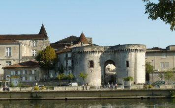 Percer les secrets de la tapisserie culturelle du Cognac Une porte historique en pierre avec deux tours rondes et une entrée voûtée se dresse au bord du fleuve à Cognac, entourée de bâtiments anciens. Les gens se promènent à pied ou à vélo le long du chemin qui longe le fleuve, se faufilant dans la tapisserie culturelle sous un ciel dégagé.