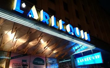 Enchanté Cinéma : Dévoilement des films français incontournables de 2022 et 2023 La photo montre l'entrée de L'Arlequin, un cinéma connu pour ses films français, avec son nom en lettres de néon bleu vif. La marquise brille au-dessus des affiches de films de 2022 2023, et un panneau bleu "L'Arlequin" est accroché près de l'extérieur chaleureusement éclairé.