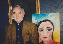 Remembering Jenny Batlay: The Luminary Who Painted Stars and Wrote with Joy Charles Aznavour en blazer et chemise noire, debout à côté d'un portrait sur un chevalet. Un autoportrait de Jenny Batlay. Le fond est bleu ciel avec une lumière dorée. Jenny porte un haut rouge et blanc et regarde vers le bas et légèrement vers sa gauche avec de grands yeux vert foncé expressifs.