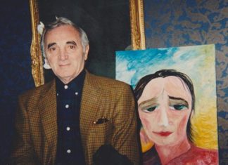 Remembering Jenny Batlay: The Luminary Who Painted Stars and Wrote with Joy Charles Aznavour en blazer et chemise noire, debout à côté d'un portrait sur un chevalet. Un autoportrait de Jenny Batlay. Le fond est bleu ciel avec une lumière dorée. Jenny porte un haut rouge et blanc et regarde vers le bas et légèrement vers sa gauche avec de grands yeux vert foncé expressifs.