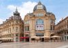 Enchanté Cinéma: Unveiling the Must-See French Films of 2024 and 2025 A wide, high-angle view of the historic Gaumont cinema building at the Place de la Comédie in Montpellier, France. The ornate, sand-colored stone architecture features classical domes and grand arched windows. The foreground is a vast, empty pedestrian plaza under a bright, partly cloudy blue sky, with outdoor seating area.