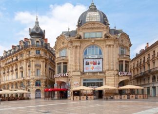 Enchanté Cinéma: Unveiling the Must-See French Films of 2024 and 2025 A wide, high-angle view of the historic Gaumont cinema building at the Place de la Comédie in Montpellier, France. The ornate, sand-colored stone architecture features classical domes and grand arched windows. The foreground is a vast, empty pedestrian plaza under a bright, partly cloudy blue sky, with outdoor seating area.
