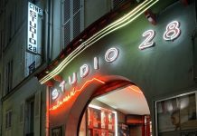 Enchanté 2026: The Essential New Releases & Production Buzz A vibrant nighttime shot of the iconic Studio 28 cinema in Montmartre, Paris. The entrance is illuminated by glowing white and orange neon signs against a dark facade. Red-carpeted steps lead up into a brightly lit lobby adorned with movie posters, situated on a narrow, atmospheric cobblestone street.