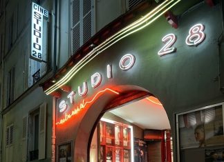 Enchanté 2026: The Essential New Releases & Production Buzz A vibrant nighttime shot of the iconic Studio 28 cinema in Montmartre, Paris. The entrance is illuminated by glowing white and orange neon signs against a dark facade. Red-carpeted steps lead up into a brightly lit lobby adorned with movie posters, situated on a narrow, atmospheric cobblestone street.